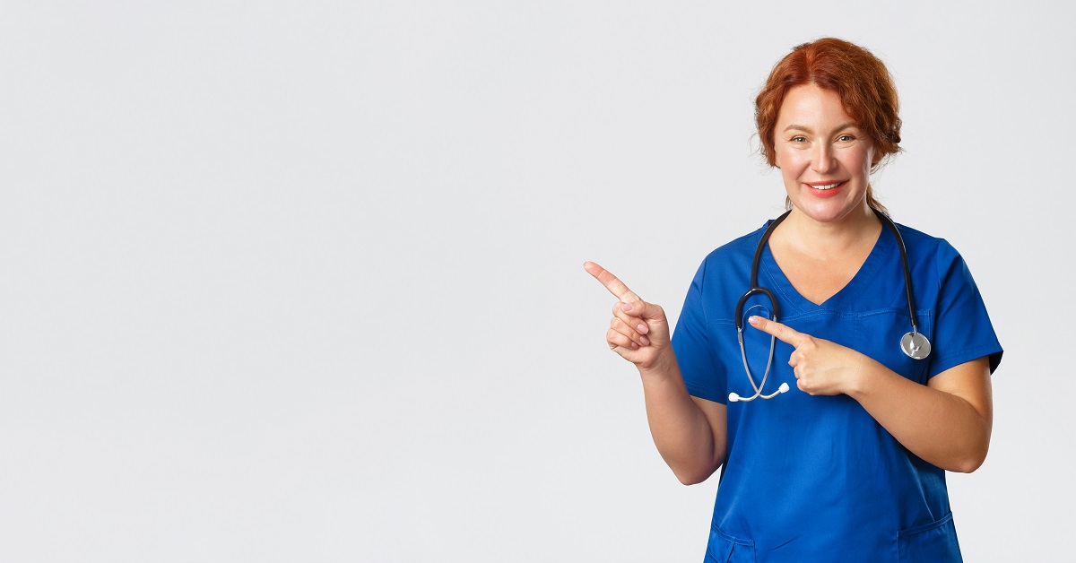Medicine, healthcare and coronavirus concept. Portrait of pleasant female doctor, redhead nurse in blue scrubs looking upbeat, showing clinic banner, hospital advertisement, pointing finger left.