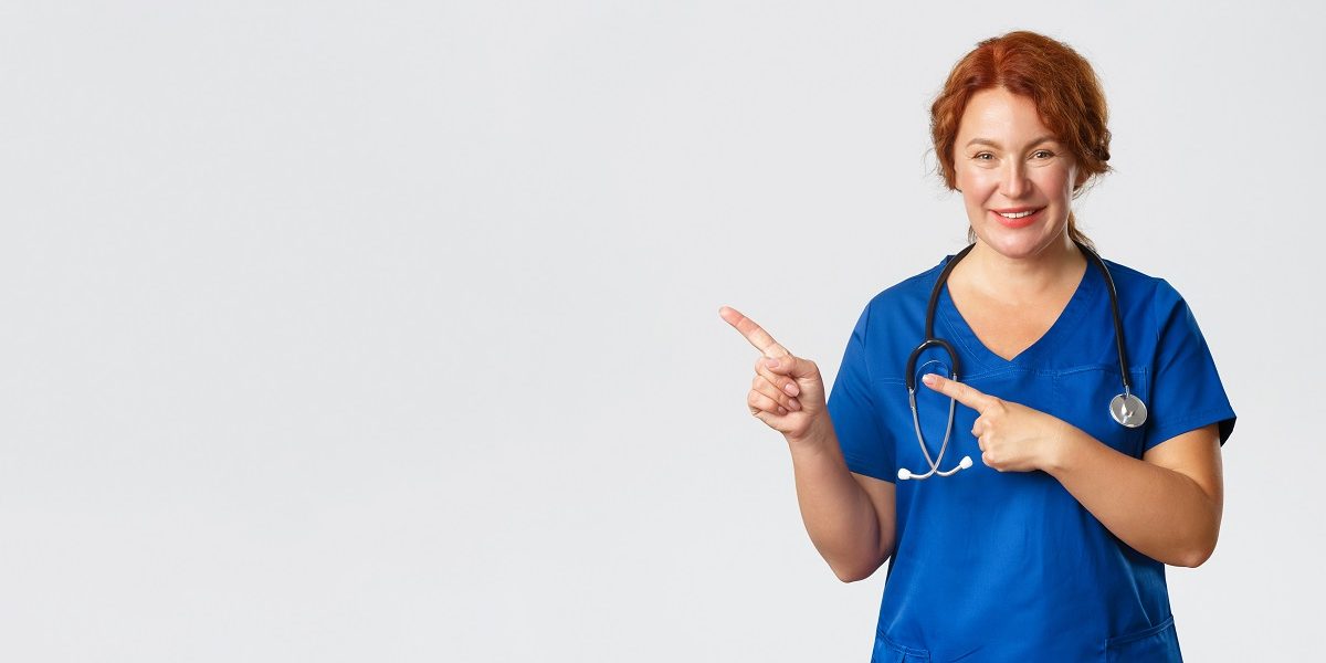 Medicine, healthcare and coronavirus concept. Portrait of pleasant female doctor, redhead nurse in blue scrubs looking upbeat, showing clinic banner, hospital advertisement, pointing finger left.